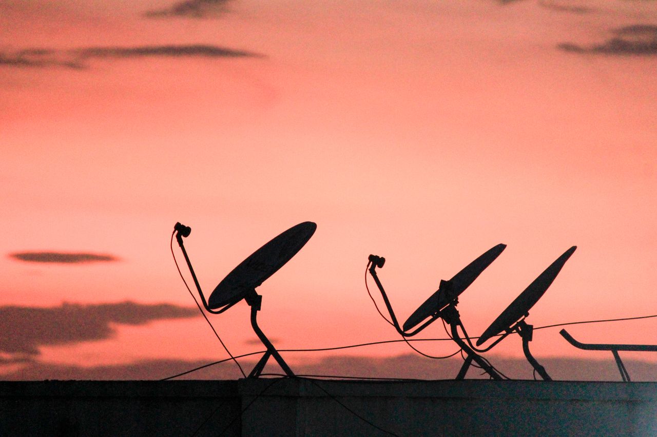 who-we-are A row of satellite dishes silhouetted against a vibrant orange sunset sky on a rooftop.